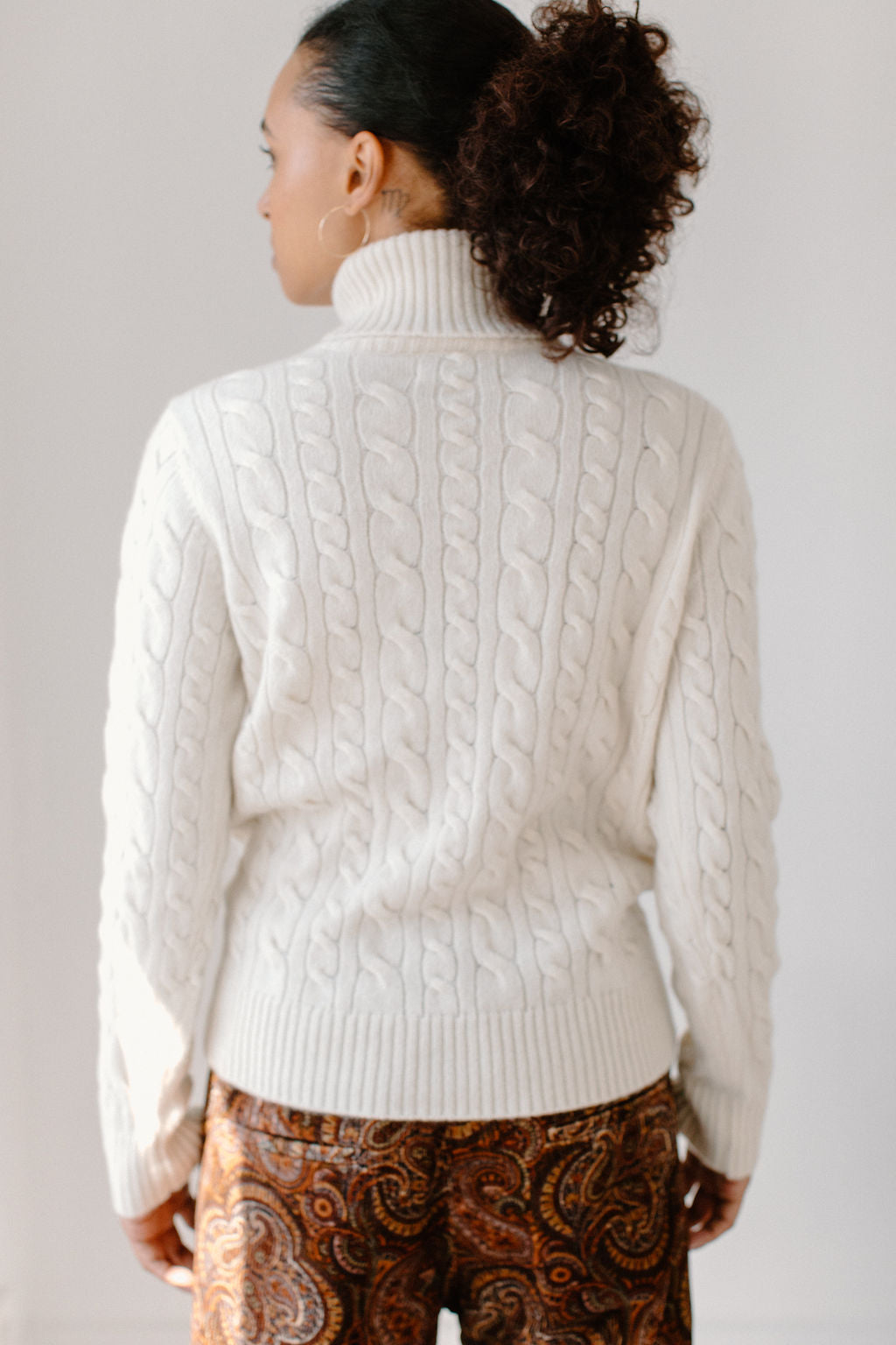 Back view of model wearing the Cream Cable-Knit Turtleneck sweater paired with paisley patterned pants, showcasing the sweater&#39;s texture.