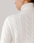 Close up of model wearing the Cream, Cable-Knit Turtleneck sweater against a white background to show the sweater's design and texture.