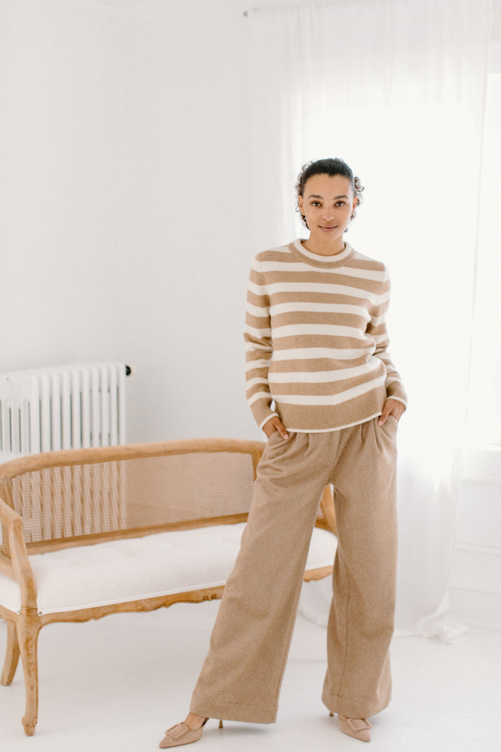 Model wearing the camel/cream Classic Striped Cashmere Crew, paired with wide-leg camel trousers and beige flats, in a minimalist room.