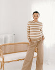 Model wearing the camel/cream Classic Striped Cashmere Crew, paired with wide-leg camel trousers and beige flats, in a minimalist room.