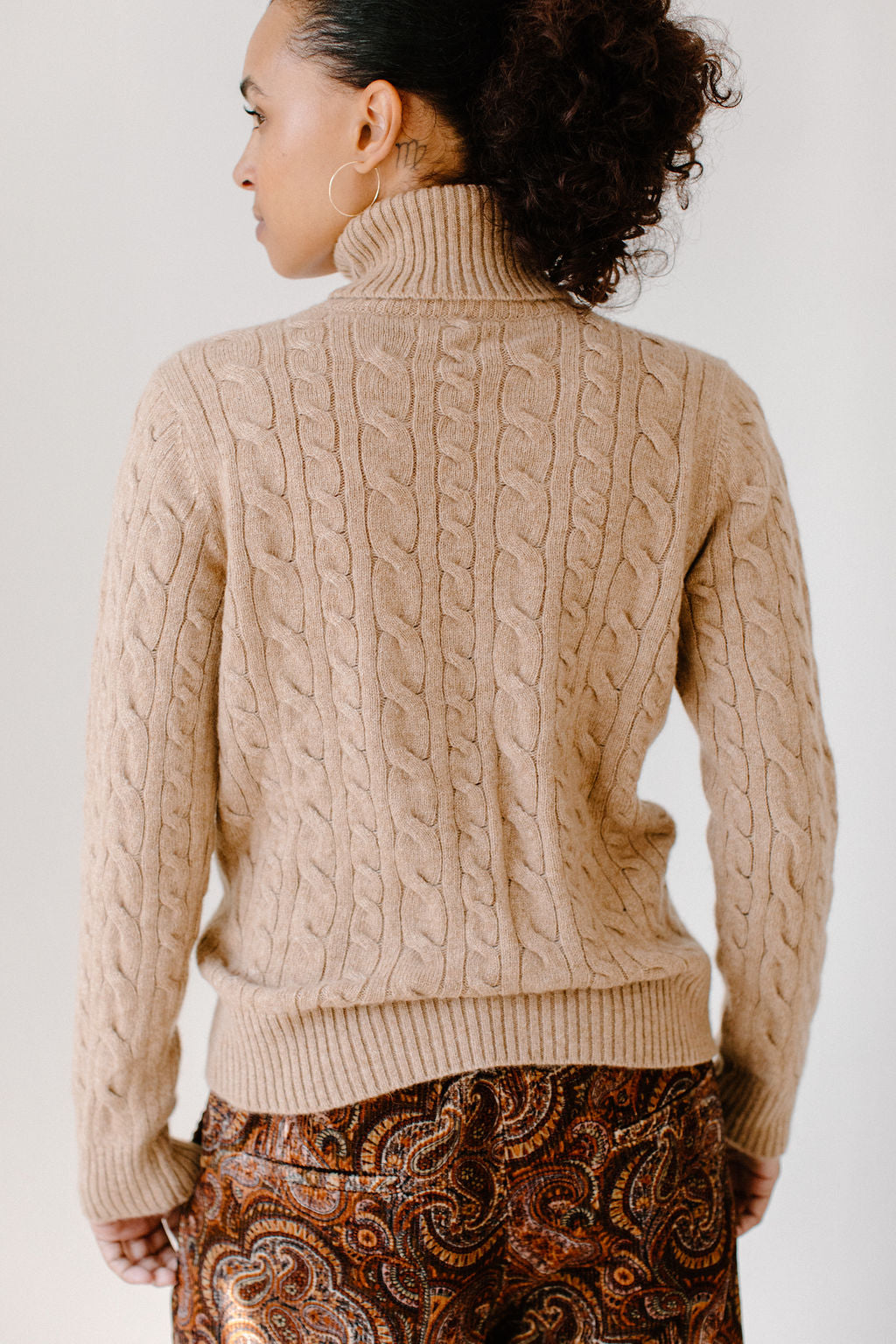 Back view of model wearing the Camel Cable-Knit Turtleneck sweater paired with paisley patterned pants, showcasing the sweater's texture.