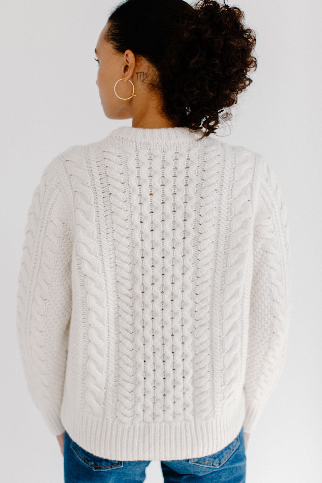 Back view of model wearing the Cream cable knit Fisherman Crewneck, paired with jeans and silver hoop earrings against a white background.