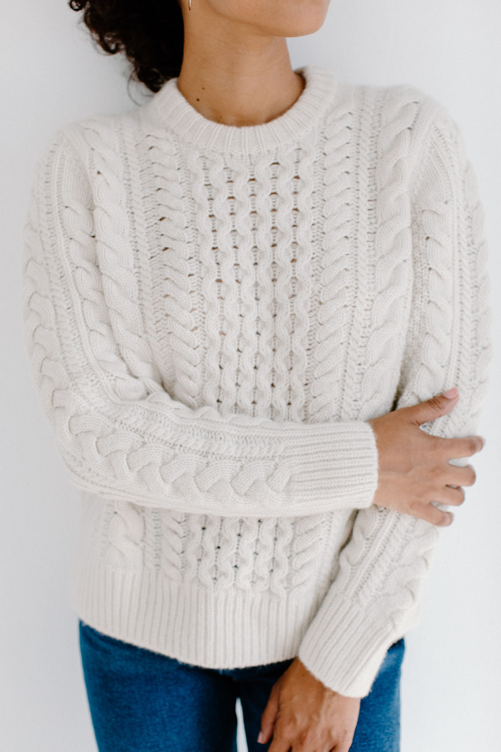 Model wearing the Cream chunky Aran cable knit Fisherman Crewneck sweater, paired with jeans in front of a white background.