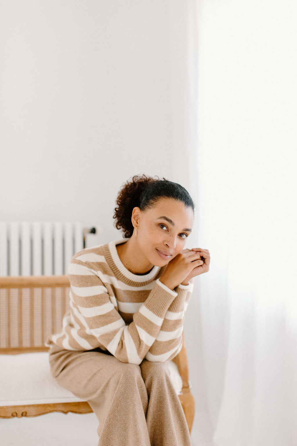 Model sitting on a chair wearing the camel/cream Classic Striped Cashmere Crew paired with camel wide-leg trousers, in a minimalist room.