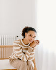Model sitting on a chair wearing the camel/cream Classic Striped Cashmere Crew paired with camel wide-leg trousers, in a minimalist room.