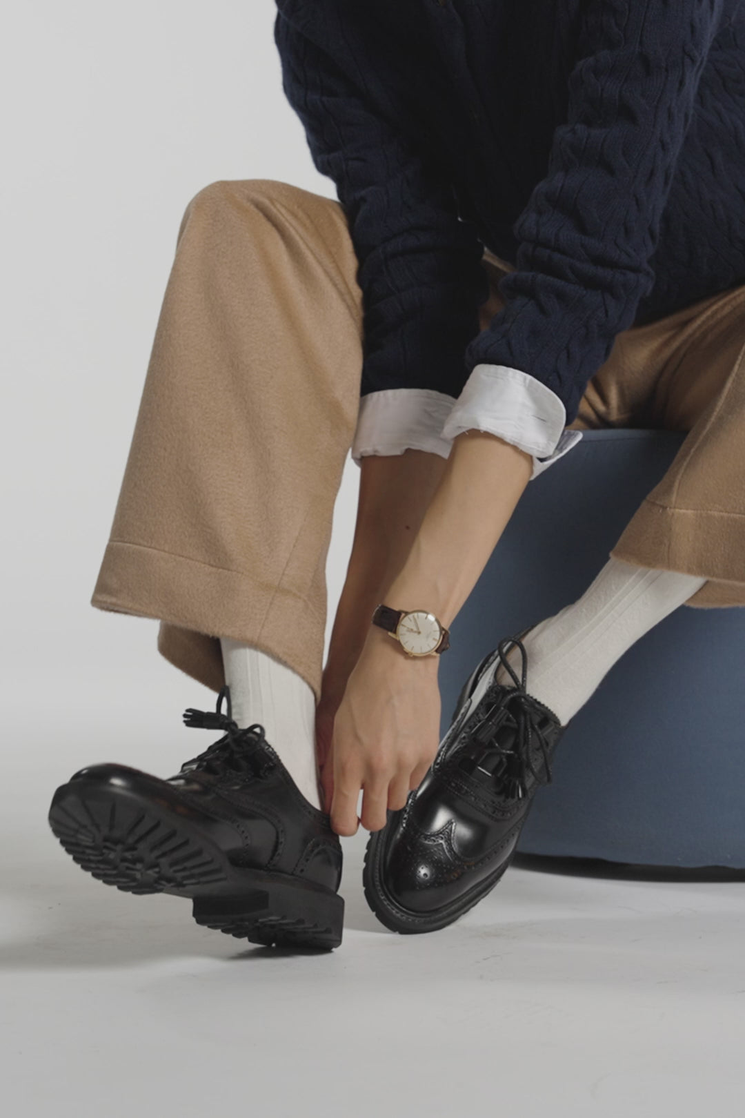 Woman putting on a black shoe wearing brown pants and white ankle socks in a studio