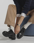 Woman putting on a black shoe wearing brown pants and white ankle socks in a studio