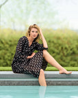 Woman in a black polka dot dress sitting by a pool with greenery in the background