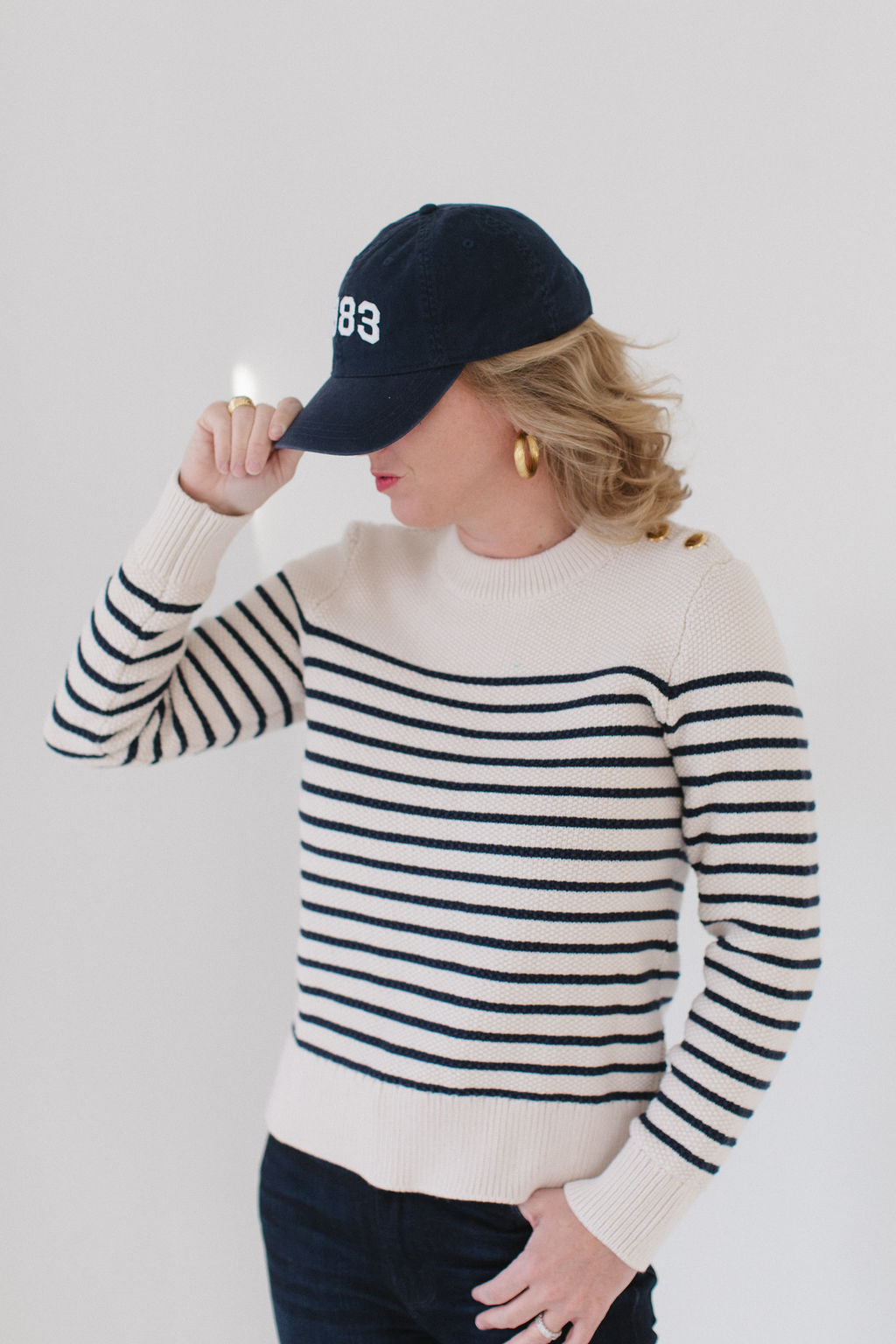 EL wearing the Perfect Striped Crewneck in cream with navy mariner stripes, paired with a '1983' cap, gold hoop earrings, and dark pants.