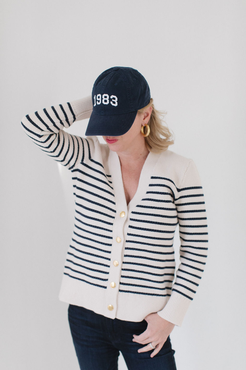 EL wearing The Perfect Striped Cardigan in cream with navy mariner stripes, paired with a '1983' cap, gold hoop earrings, and dark jeans.