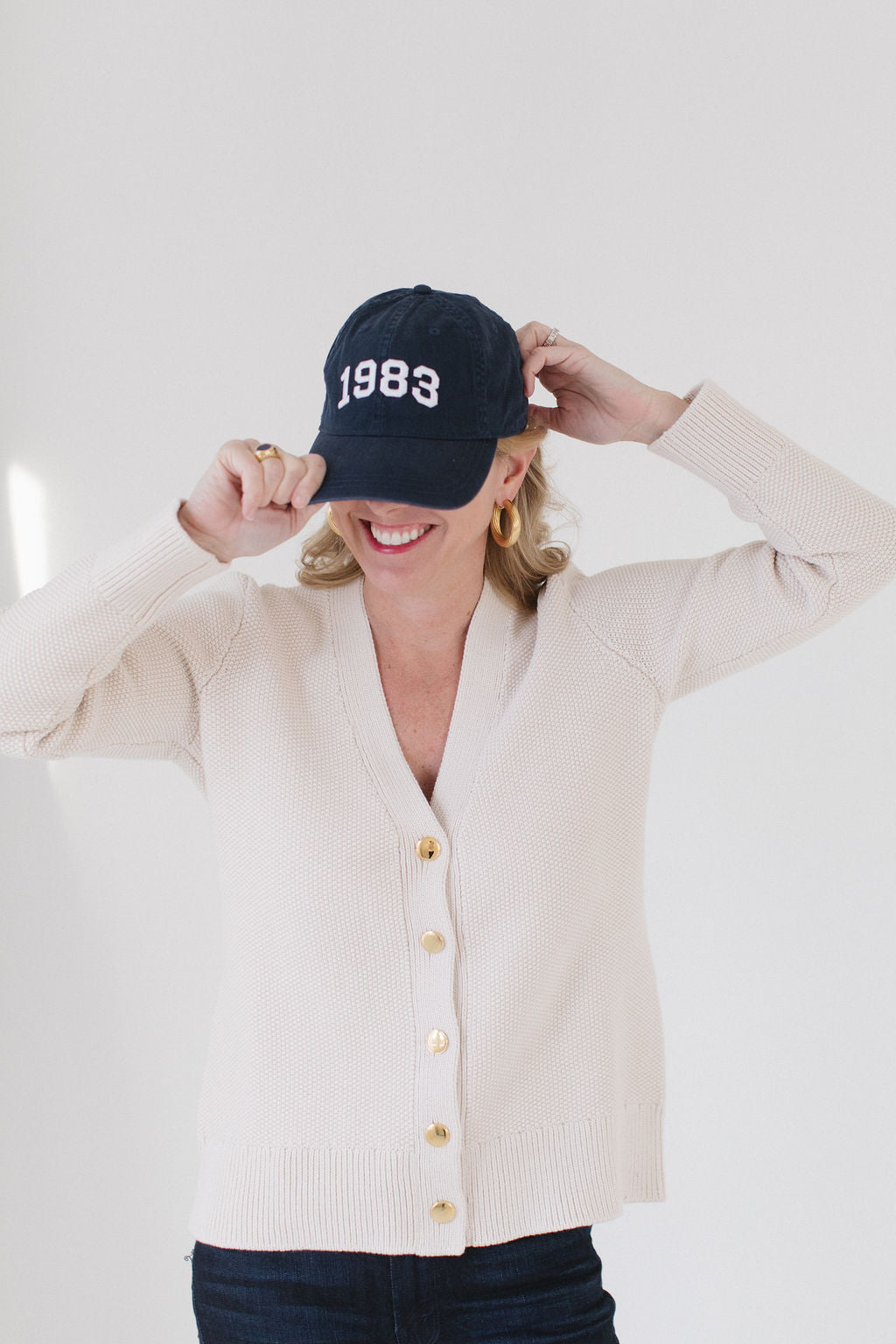 Eleanor Leftwich wearing The Perfect Cardigan in cream paired with a navy '1983' cap and gold hoop earrings against a white background.
