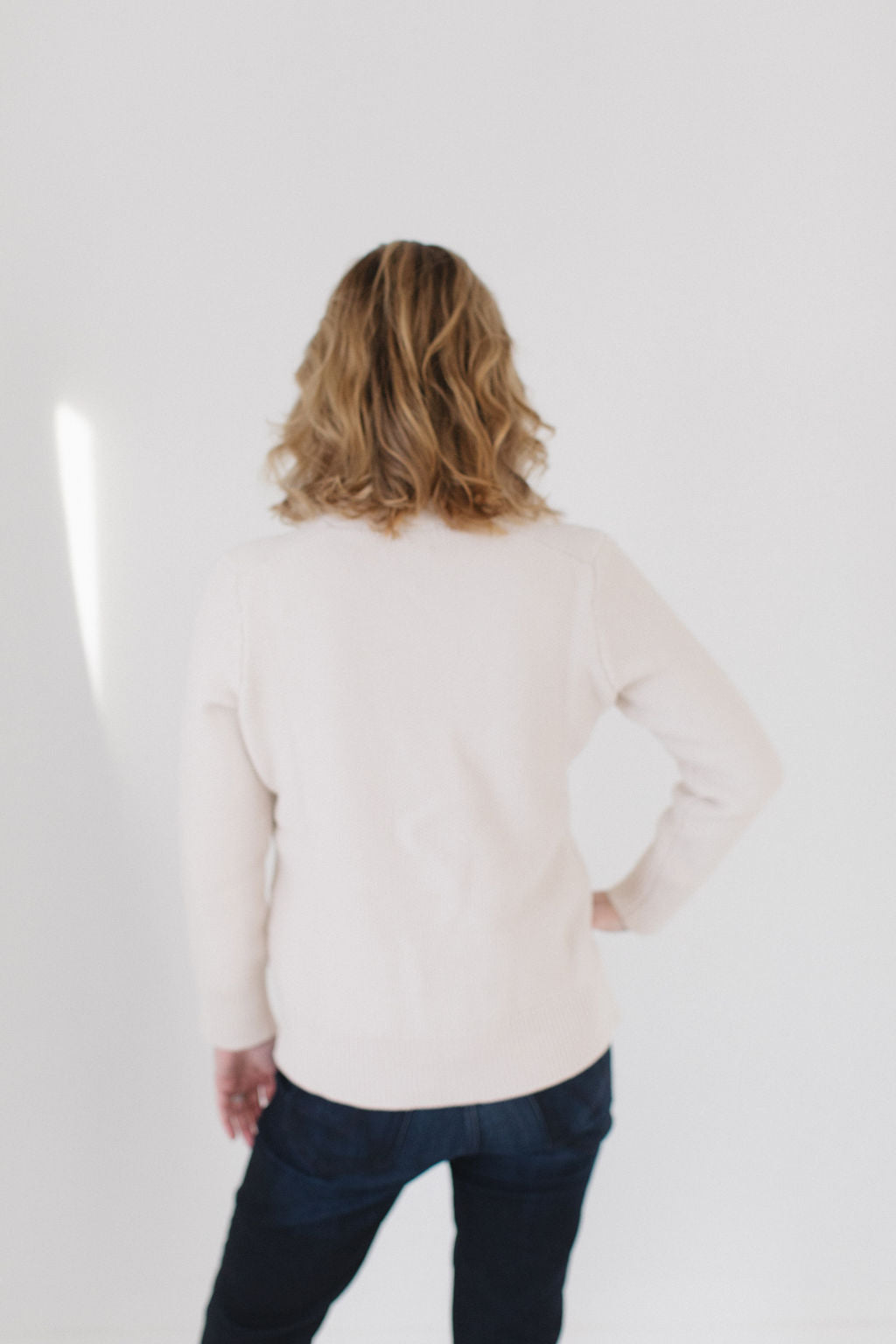 Back view of Eleanor Leftwich wearing The Perfect Cardigan in cream paired with dark denim jeans, facing a white background.