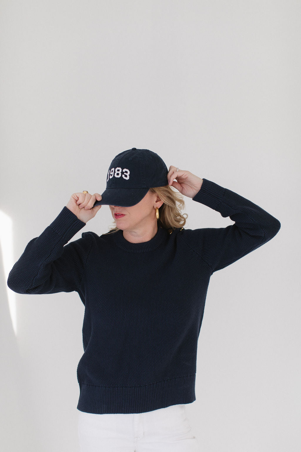 EL wearing the Navy Perfect Crewneck, paired with a '1983' cap, gold hoop earrings, and white pants, adjusting the cap with both hands.