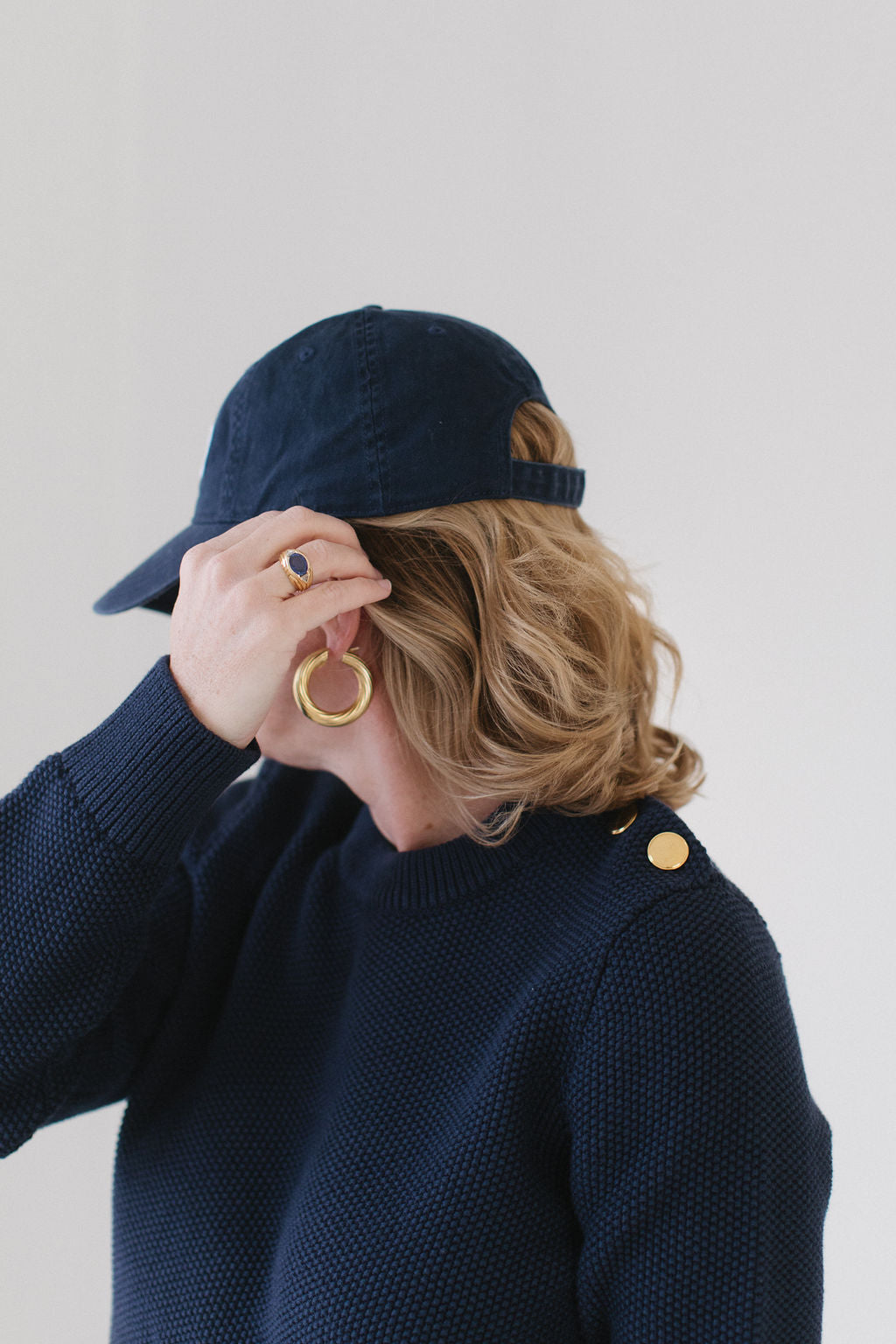 Close-up of EL in the Navy Perfect Crewneck with gold buttons on the shoulder, paired with a navy cap, gold hoop earrings, and a gemstone ring.