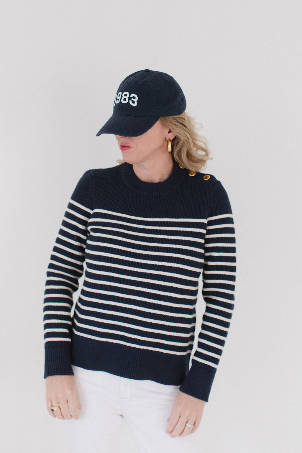 EL wearing the Perfect Striped Crewneck in navy with cream mariner stripes, paired with a '1983' cap, gold hoop earrings, and white pants.