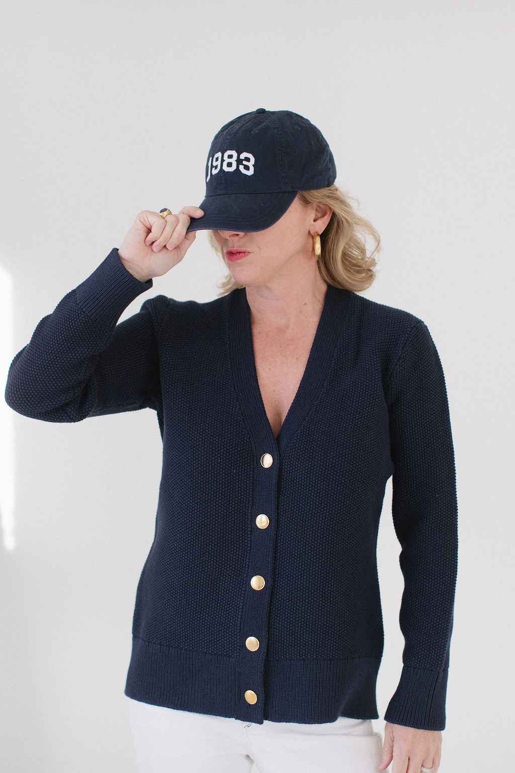 Eleanor Leftwich wearing The Perfect Cardigan in navy with gold buttons, paired with a '1983' cap, gold hoop earrings, and white pants.